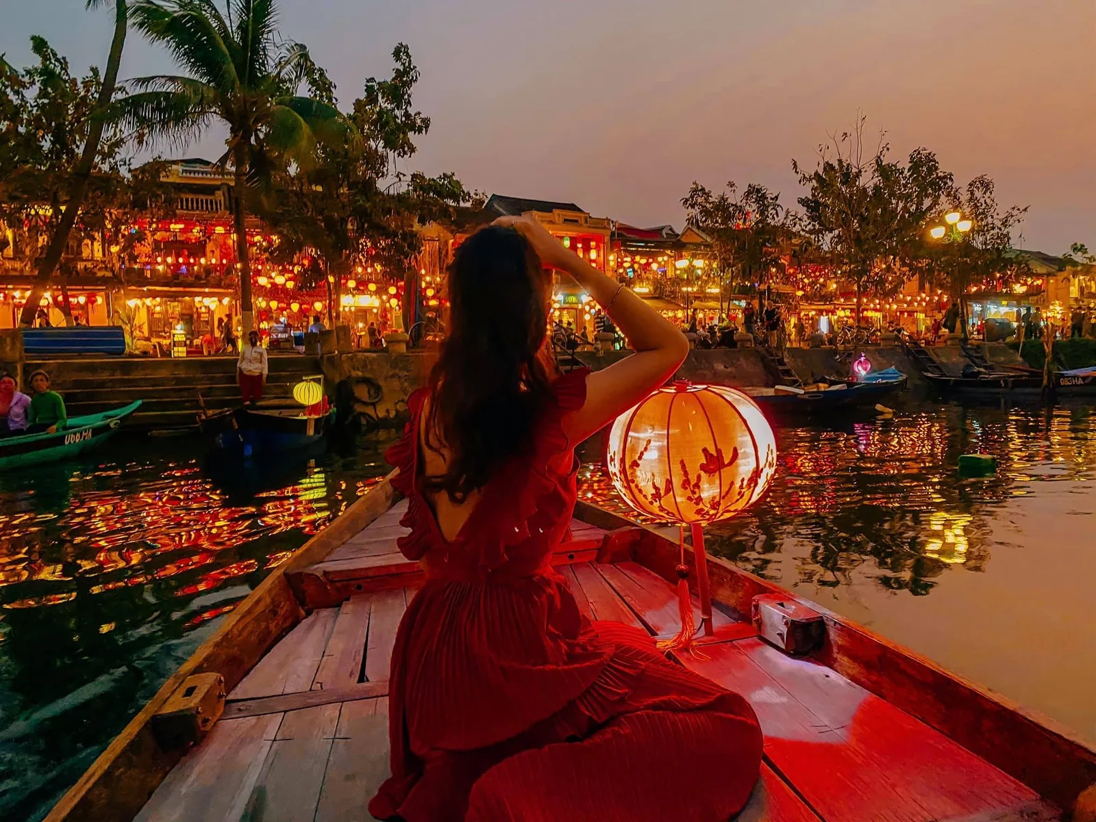 Người phụ nữ mặc váy đỏ ngồi trên thuyền bên đèn lồng, ngắm phố cổ Hội An lấp lánh ánh đèn lúc chạng vạng.
